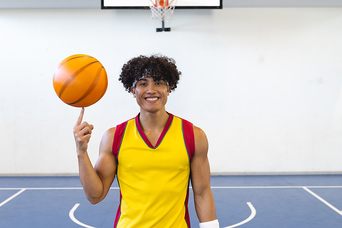Young biracial man spins a basketball on his finger at a gym. His cheerful expression and athletic attire suggest a passion for the sport.