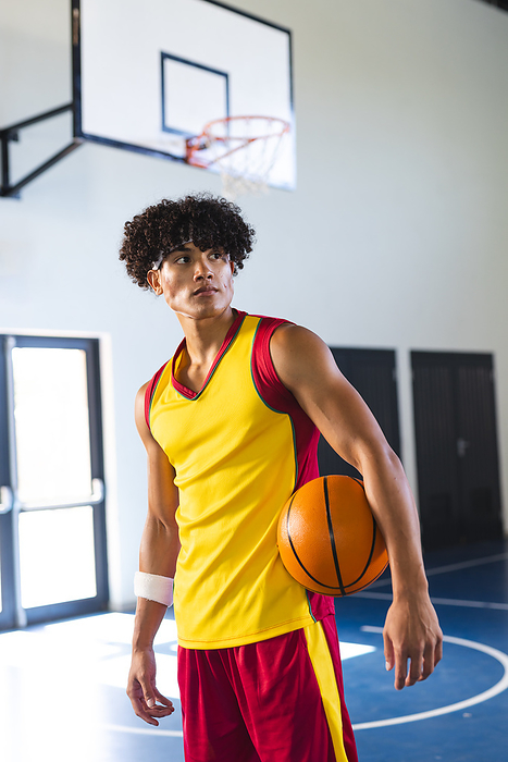 Young biracial man holds a basketball in a gym. His focused gaze suggests determination and readiness for the game.