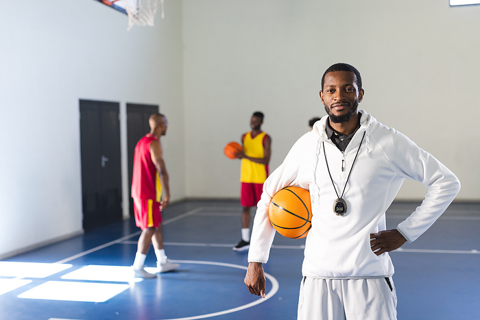 African American coach poses confidently on a basketball court. Teammates practice in the background, highlighting a sports and fitness theme.