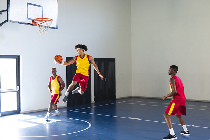 Young African American man jumps for a basketball shot in a gym. Teammates watch eagerly as the game unfolds indoors.