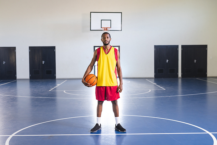 African American man stands confidently on a basketball court. His athletic attire suggests he is ready for a game at the school gym.