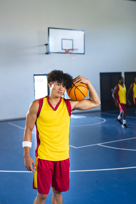 Young biracial man poses with a basketball in a gym, with copy space. His confident stance suggests he's ready for a game at the indoor court.