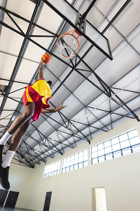 African American man scores during a basketball game in an indoor court with copy space. His athletic prowess is showcased as he leaps towards the hoop.
