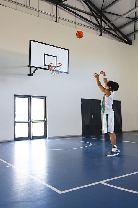 Young biracial man practices shooting a basketball in a gym with copy space. Athletic student perfects his skills on the indoor court at school.