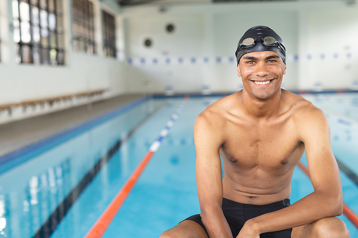 Young biracial male athlete swimmer smiles at poolside, with copy space. His cheerful expression brightens the indoor swimming facility.