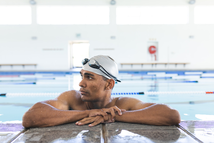Young biracial male athlete swimmer rests at the poolside, with copy space. He's taking a break during training at an indoor swimming facility.