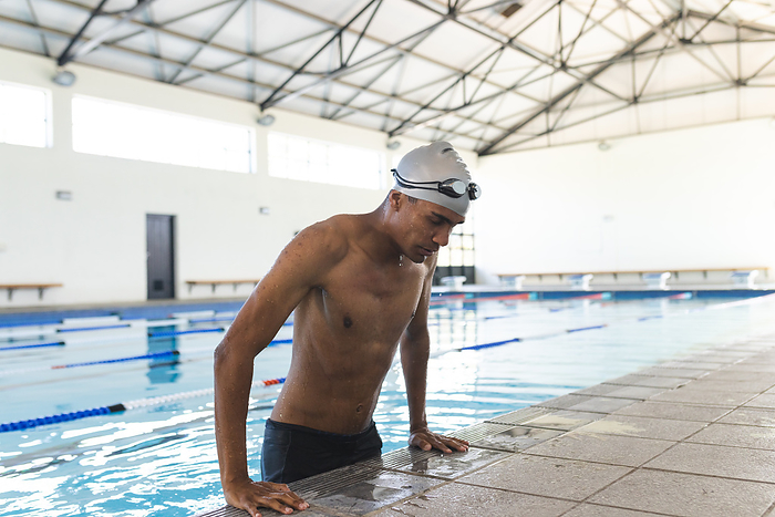 Young biracial male athlete swimmer rests at the poolside after swimming. Indoor pool setting captures the athlete's moment of recovery.