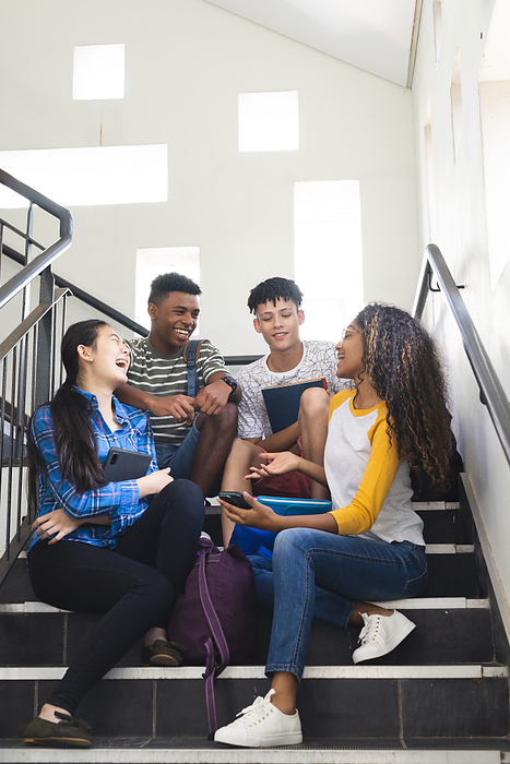 Teenage biracial boys and a teenage girl chat on high school stairs. Their laughter and conversation capture a moment of youthful camaraderie in a high school setting.