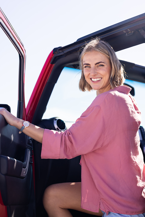 Young Caucasian woman smiles while entering a car on a road trip. Her cheerful expression suggests excitement for a new adventure.