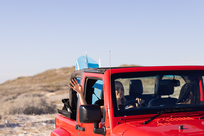 Young African American woman and young Caucasian woman enjoy a sunny beach drive on a road trip. Smiling faces and surfboards hint at an adventurous day outdoors.