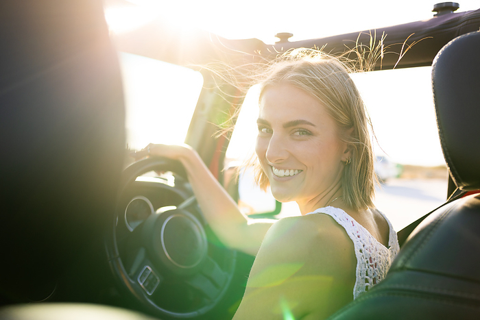 Young Caucasian woman smiles while driving on a road trip. Her joyful expression suggests a sense of freedom during a road trip or commute.