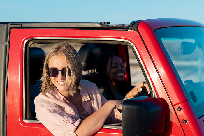 Young Caucasian woman and African American woman enjoy a car ride on a road trip. Smiles radiate as they share a moment on a sunny outdoor adventure.