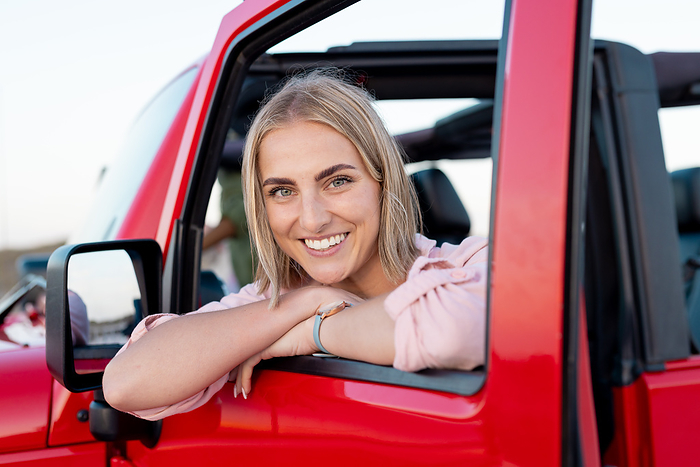 Young Caucasian woman smiles from a red car on a road trip. Her cheerful expression suggests a sense of freedom and joy during an outdoor adventure.