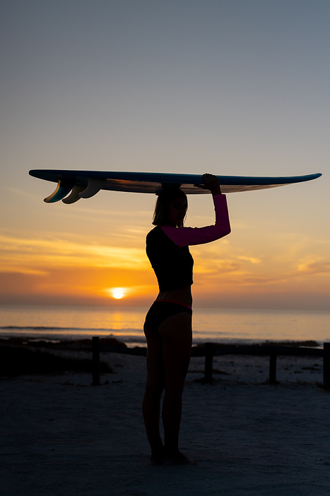 Silhouetted young Caucasian woman holds a surfboard at sunset, with copy space. She stands on the beach, capturing the essence of outdoor adventure and leisure.