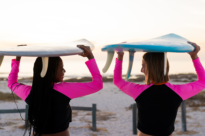 Young African American woman and young Caucasian woman carry surfboards on a beach. They enjoy a sunset surf session, highlighting an active outdoor lifestyle.