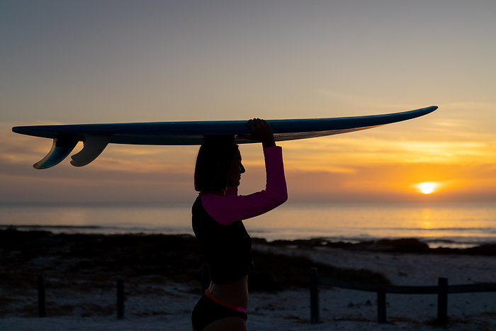 Silhouette of a young Caucasian woman carrying a surfboard at sunset, with copy space. She's at the beach, ready for an evening surf session.