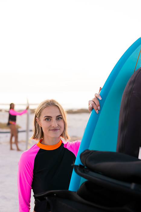 Young Caucasian woman holds a surfboard on the beach, with copy space. She's ready for a surfing session at sunset, capturing the essence of outdoor adventure.