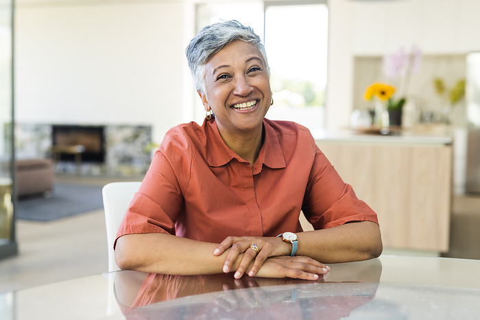 Senior smiling biracial woman seated at a table indoors on a video call. Her cheerful expression brightens the home setting.