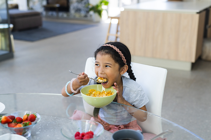Biracial girl enjoys her breakfast at home, with copy space. She's seated at a modern dining table, starting her day with a healthy meal.