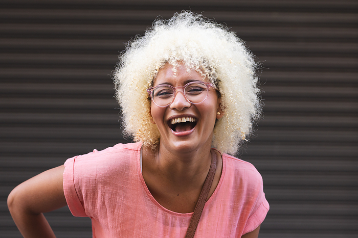 Young biracial woman laughs joyfully in the city against a striped background. Her bright smile and casual style convey a sense of genuine happiness and ease.