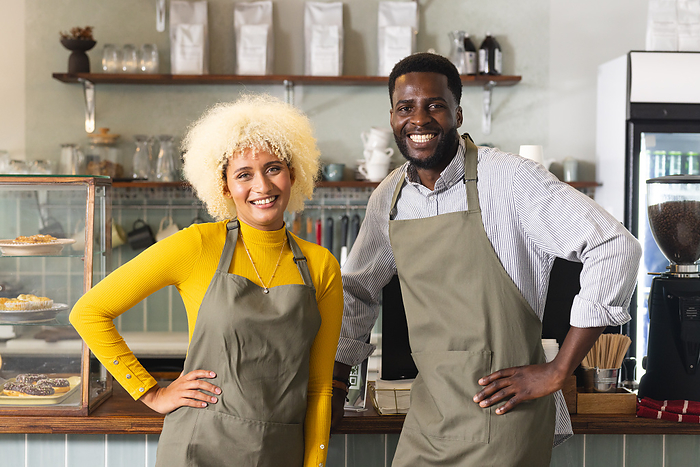 Young African American man and young Biracial woman stand in a cafe. They wear aprons, suggesting they are baristas or cafe owners, with a friendly demeanor.