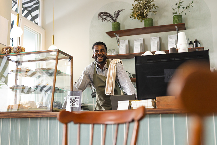 African American businessman smiles behind the cafe counter. His warm greeting invites customers into the cozy coffee shop setting.