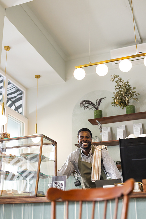 An African American male barista stands behind a cafe counter, with copy space. His welcoming smile adds a friendly atmosphere to the cozy coffee shop setting.