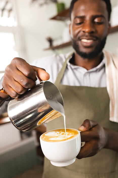 African American barista pours milk into a coffee at a cafe. His skillful preparation suggests a cozy, inviting atmosphere for coffee enthusiasts.