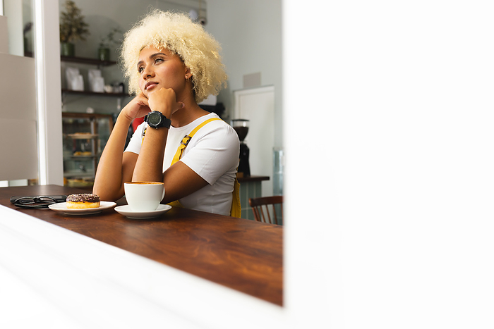 Young biracial woman enjoys a moment of contemplation at a cafe, with copy space. A cup of coffee and a donut accompany her as she takes a break in a cozy indoor setting.