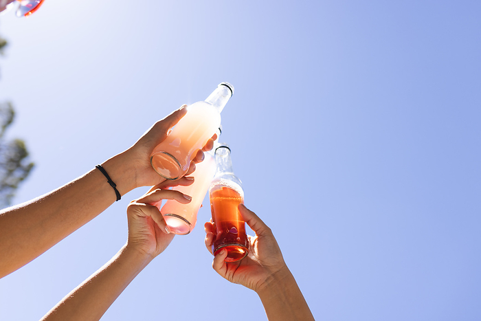 Hands clinking soda bottles against a clear blue sky. The outdoor setting suggests a casual, refreshing moment shared between friends.