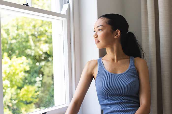 A young biracial woman gazes out a window at home. She appears contemplative as natural light illuminates the room.