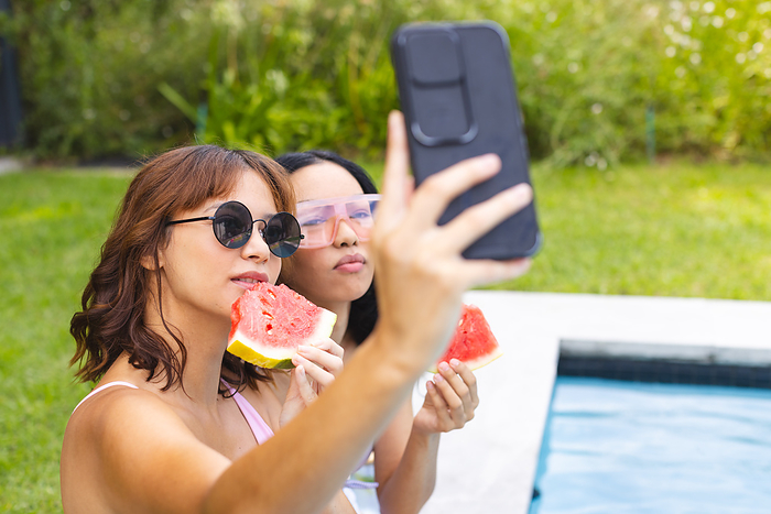 Two young biracial women enjoy a sunny poolside day, taking a selfie. They are capturing summer memories while cooling off with fresh watermelon slices.