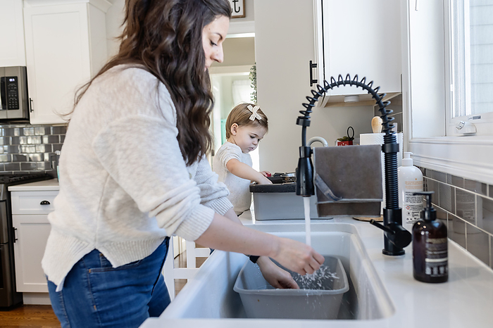 Mother Washing Dishes while Daughter pretends to wash dishes, by Cavan Images / Katie Pugliese Photography LLC
