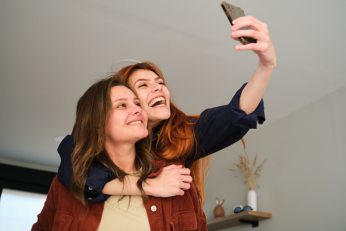 Lesbian couple laughing and taking a selfie, piggyback at home., by Cavan Images / Cristina Villar Martín