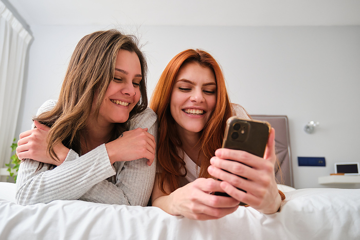 Lesbian couple laughing and using the smartphone on bed., by Cavan Images / Cristina Villar Martín
