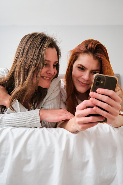 Lesbian couple smiling and using the smartphone on bed., by Cavan Images / Cristina Villar Martín