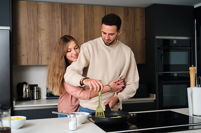 Spanish couple smiling, cooking omelette and embracing in a kitchen., by Cavan Images / Cristina Villar Martín