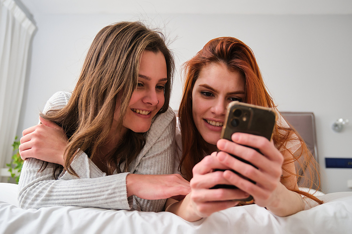 Lesbian couple using the smartphone together on bed., by Cavan Images / Cristina Villar Martín