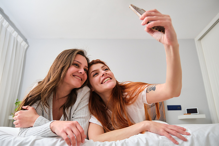 Lesbian couple smiling and taking a selfie on bed in the morning., by Cavan Images / Cristina Villar Martín