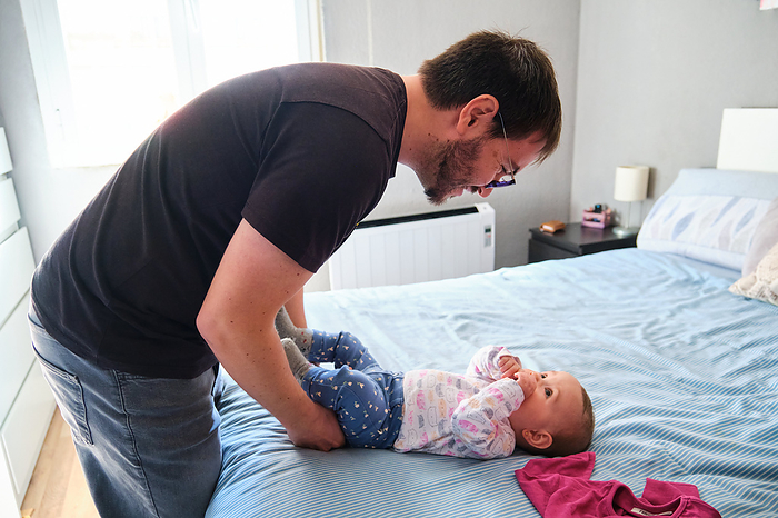 Happy father dressing his 6-Month-Old baby girl on bed., by Cavan Images / Cristina Villar Martín