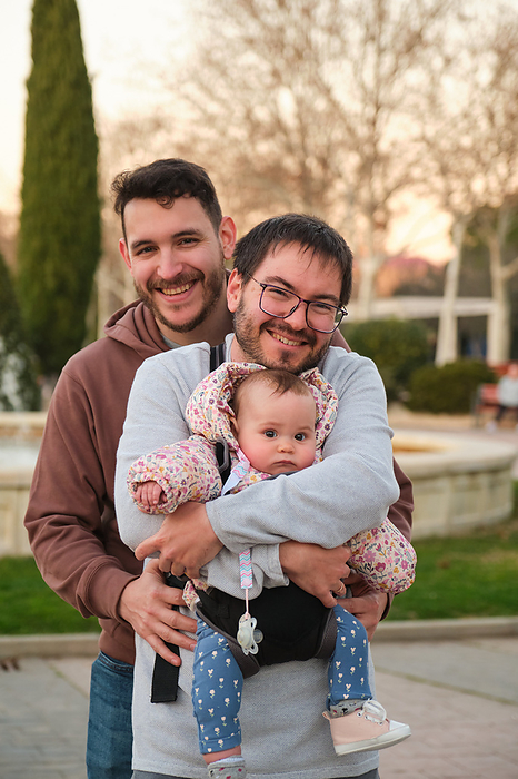 Portrait of gay happy couple with 6-Month-Old baby in a baby carrier., by Cavan Images / Cristina Villar Martín