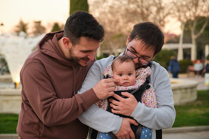 Homosexual happy couple playing with their baby in a baby carrier., by Cavan Images / Cristina Villar Martín