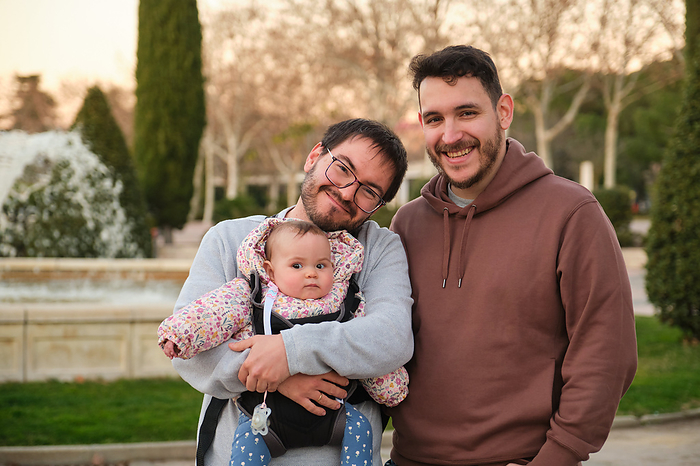Homosexual happy couple with their 6-Month-Old baby in a baby carrier., by Cavan Images / Cristina Villar Martín