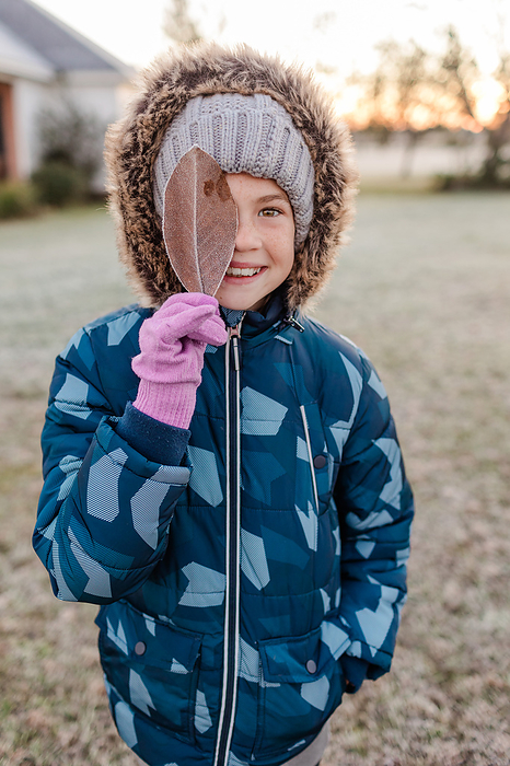 Little girl wearing a puffy winter coat holding up a frozen leaf, by Cavan Images / Captured by Colson