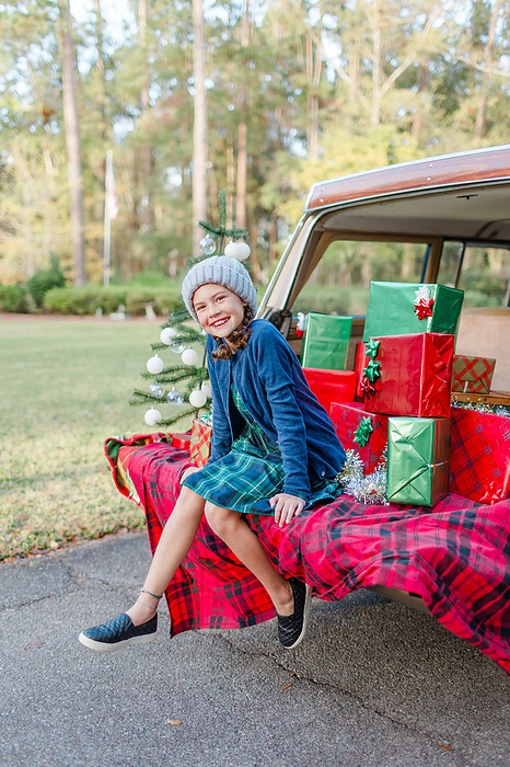 Little girl sits on the tailgate of a jeep with Christmas gifts, by Cavan Images / Captured by Colson