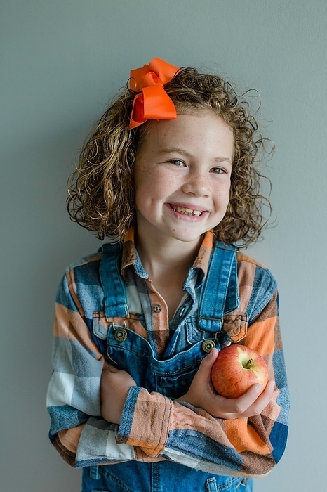 Little girl with curly hair wears overalls holding an apple, by Cavan Images / Captured by Colson
