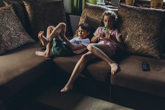 Siblings are sitting on the couch in front of the TV, by Cavan Images / Natalia Akulova