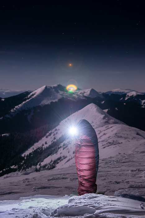 Hiker Standing Up In Sleeping Bag At Snowy Mountain Range at Night, by Cavan Images / Artur Abramiv