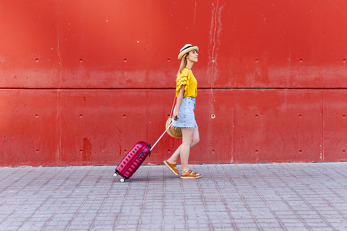 young woman walking confidently with her  suitcase against a red wall., by Cavan Images / rafa fernandez