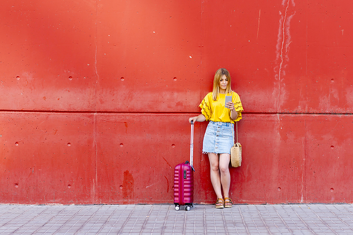 young traveler stands against a red wall, her suitcase at her side, by Cavan Images / rafa fernandez